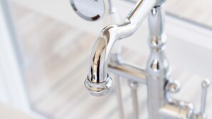 A chrome faucet with a lever handle and a separate control knob, attached to a clear glass partition.