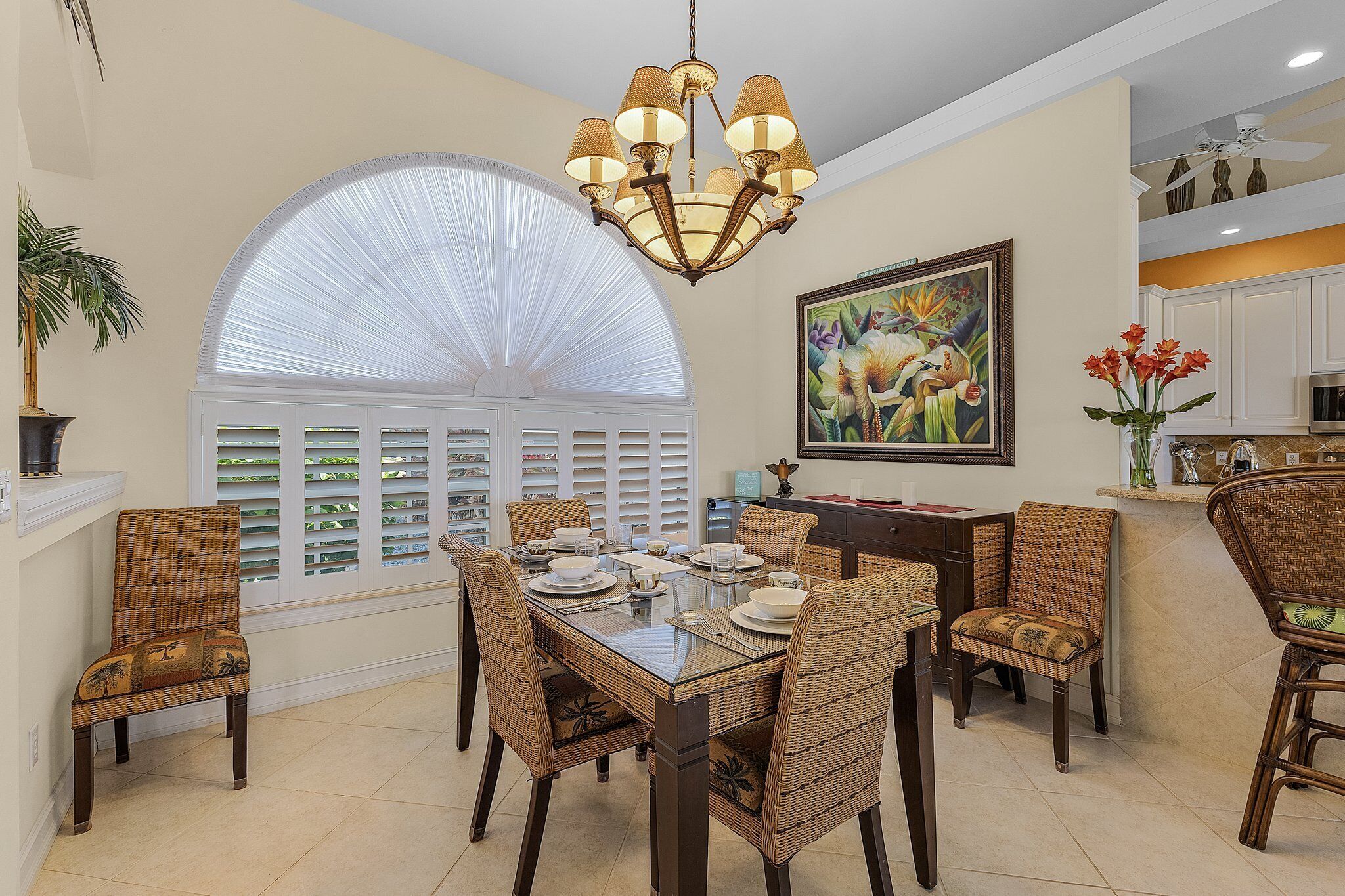 A dining area with a glass table, wicker chairs, a chandelier, a large window with blinds, and a kitchen in the background.