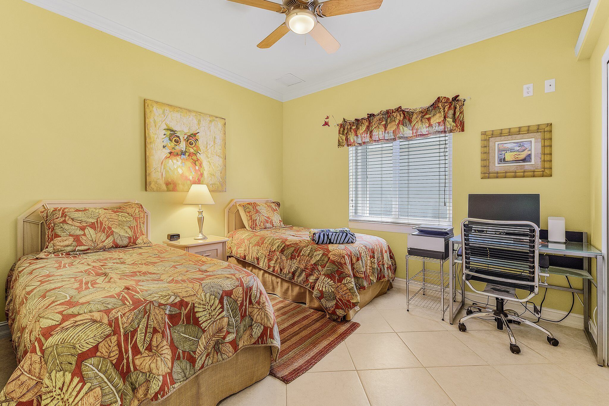 A room with two beds, a desk, a chair, a ceiling fan, and a window with blinds.