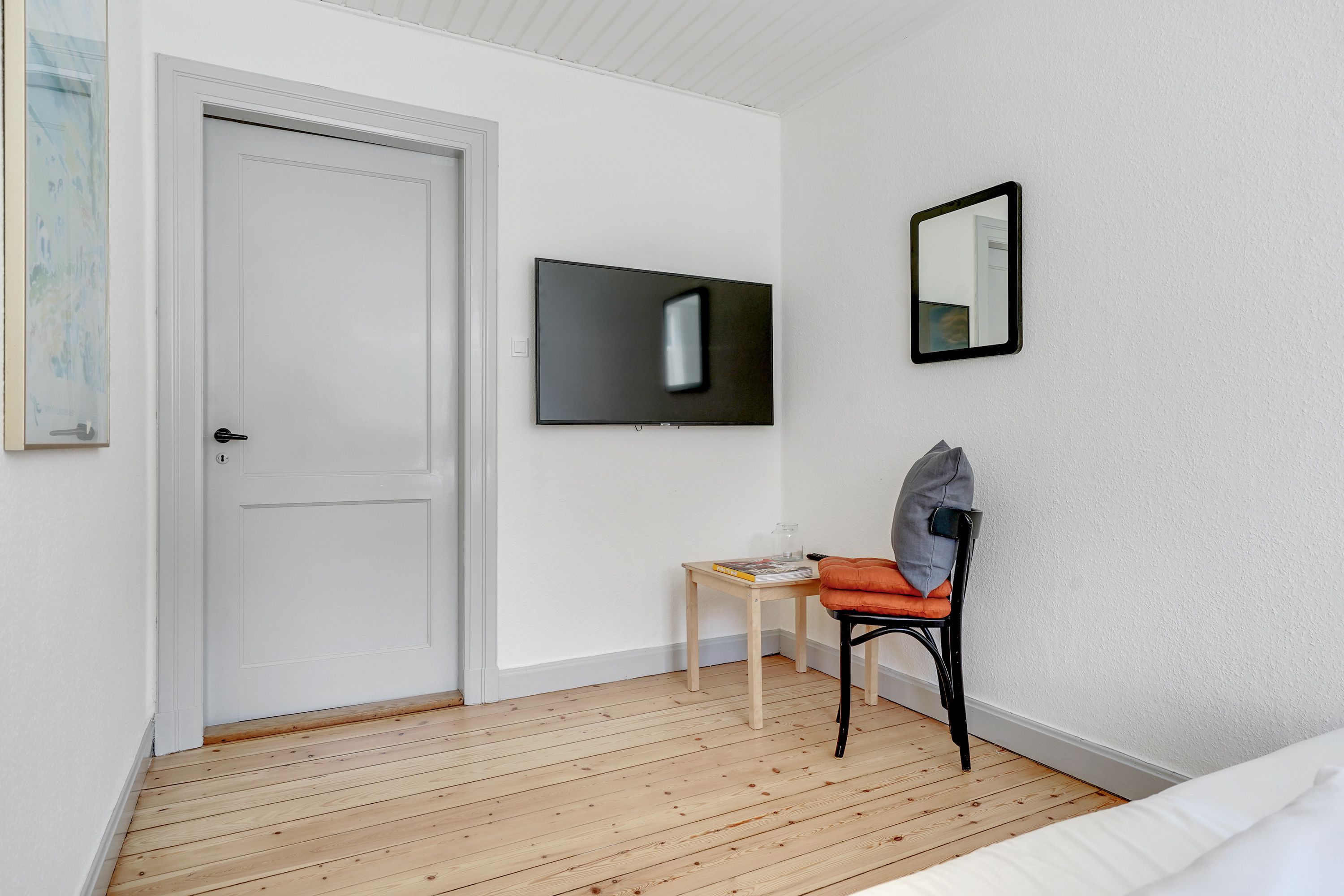 Una habitación con una puerta blanca, un televisor de pantalla plana montado en la pared, una pequeña mesa de madera con una silla y una cama con ropa blanca.