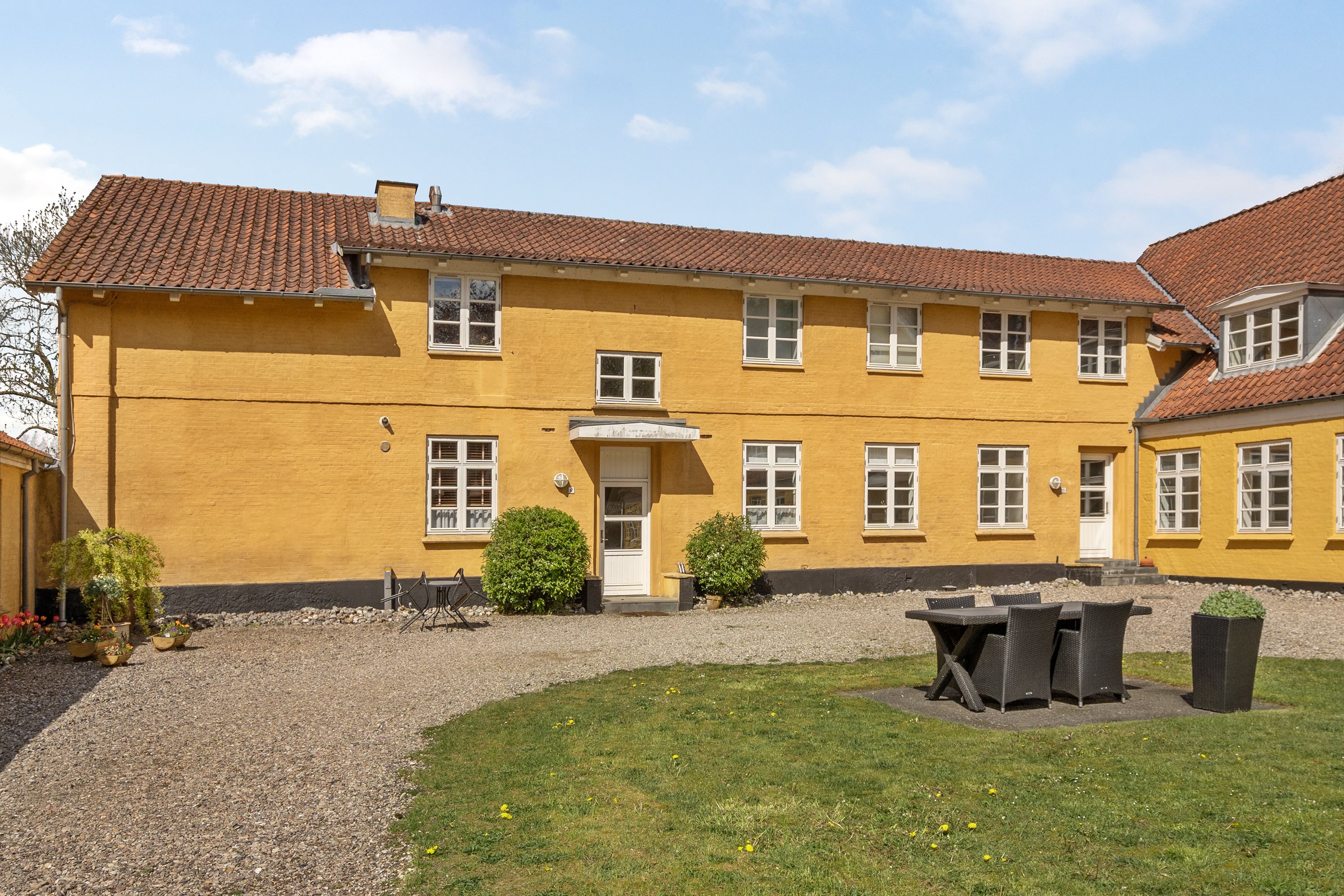 A yellow building with a tiled roof, a gravel courtyard, and outdoor seating.
