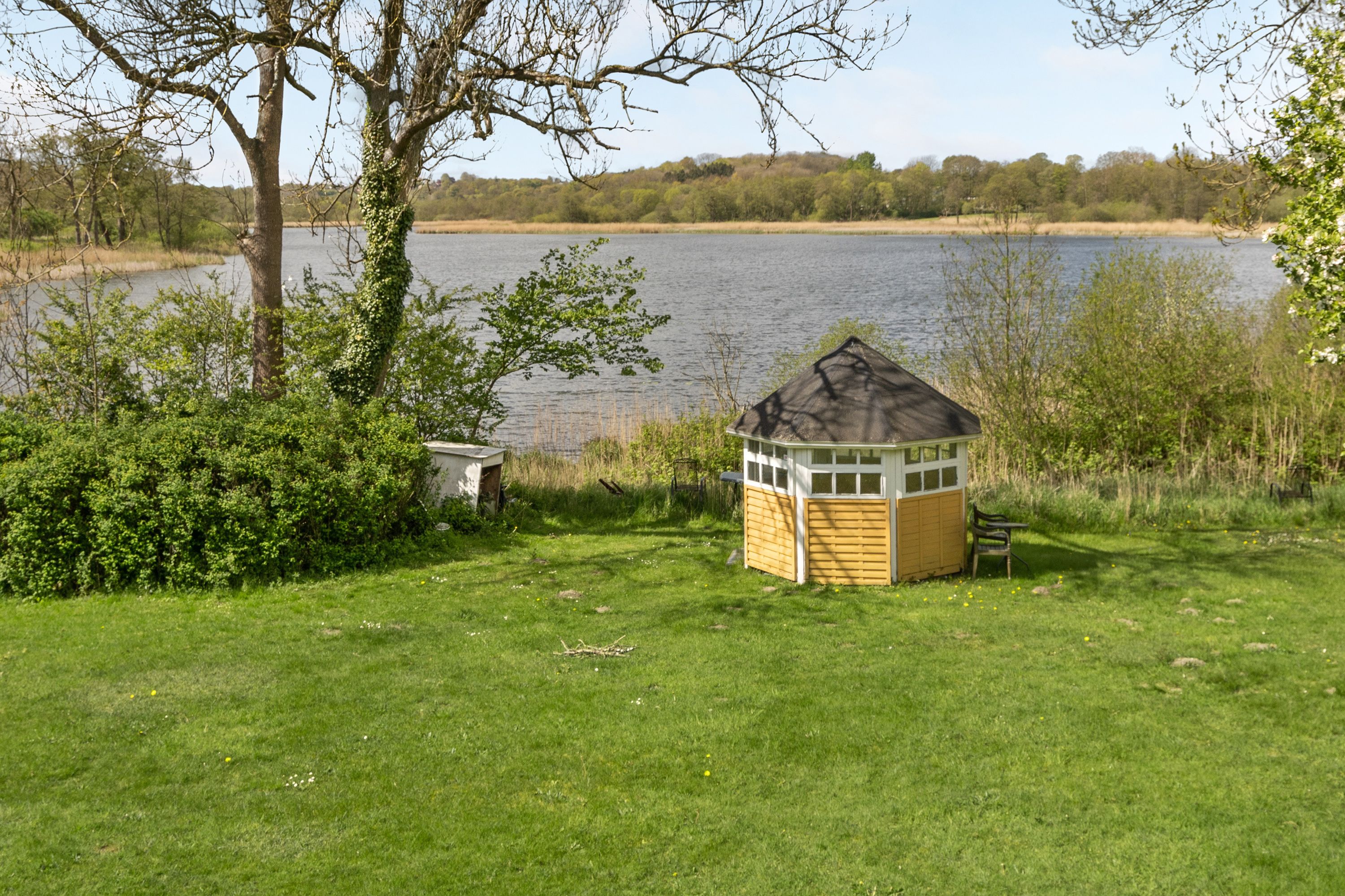 A small wooden structure with a sloped roof, a bench, and a grassy area in front of a lake.