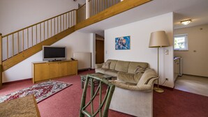 En stue med en trappe i træ, et fjernsyn på et træskab, en beige sofa, en lampe og et rødt tæppe.