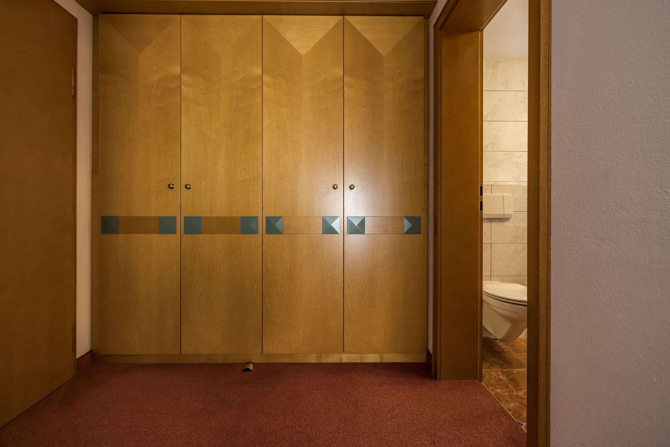 Une armoire en bois au motif géométrique, une moquette rouge et des toilettes visibles dans la pièce adjacente.