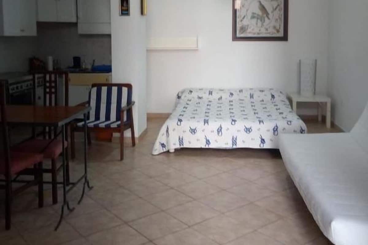 Una habitación pequeña y limpia con una cama, una silla, una mesa y un kitchenette.