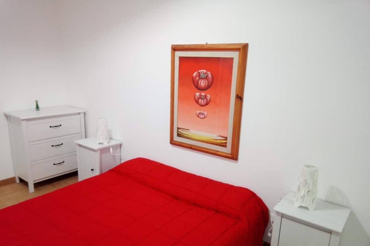 Un dormitorio con una colcha roja, un tocador blanco, una mesita de noche y un cuadro en la pared.