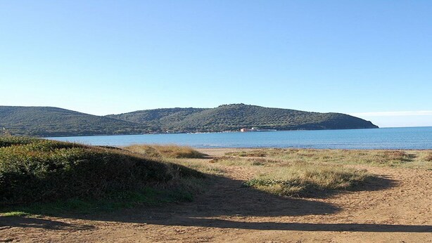 Uma praia com um caminho de terra que leva à água, rodeada por colinas e um céu claro.