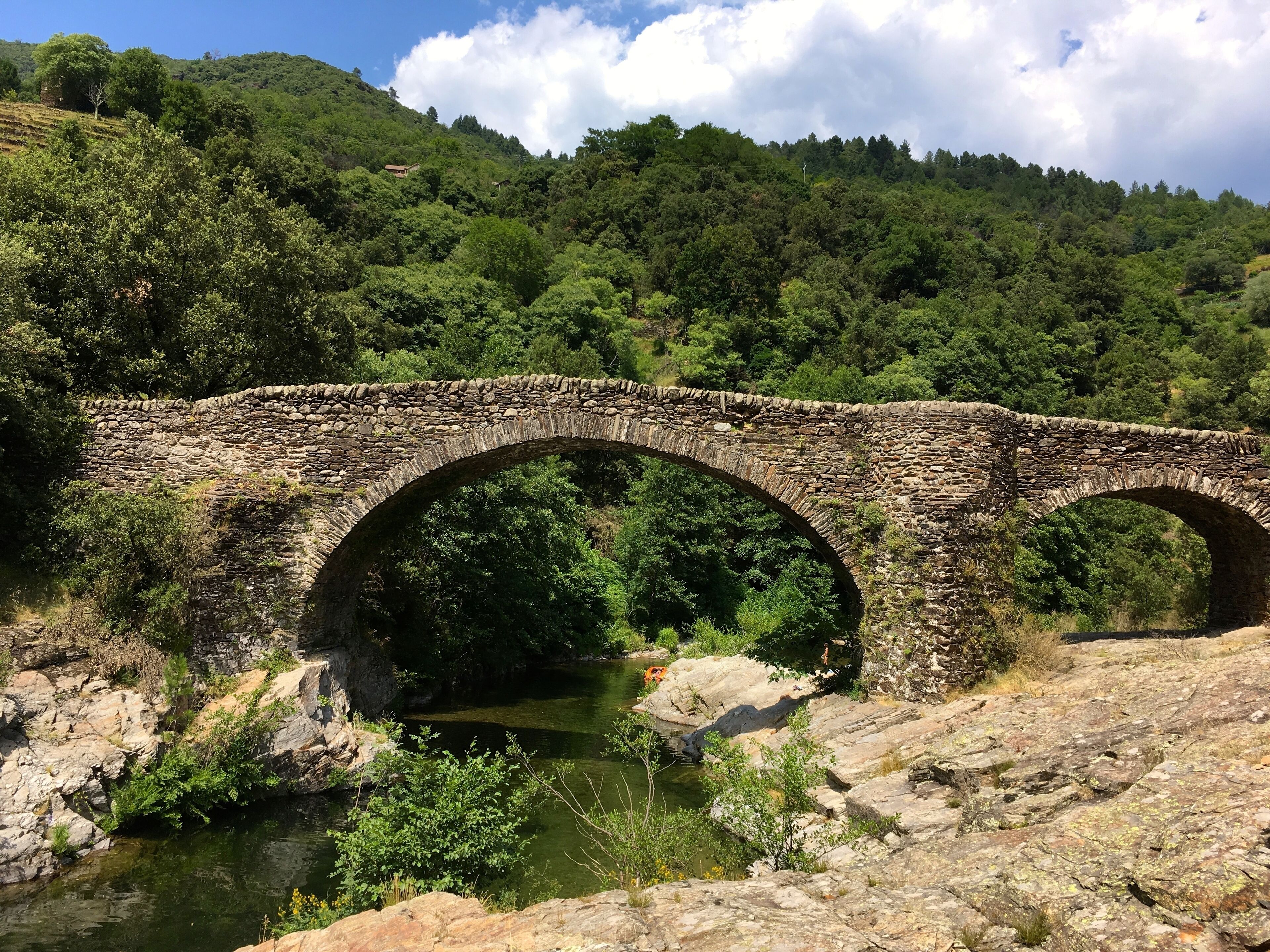 Un puente de piedra sobre un río, con vegetación exuberante y colinas al fondo.
