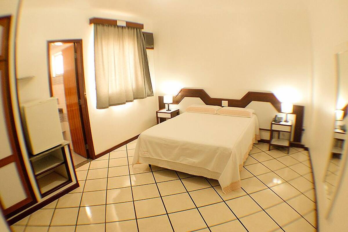 A hotel room with a bed, bedside tables, a mirror, a window with curtains, and a tiled floor.