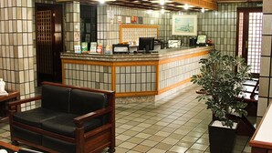 A reception area with a tiled floor, a reception desk, a black leather sofa, and a potted plant. - Shalako Hotel (Vitoria da Conquista)