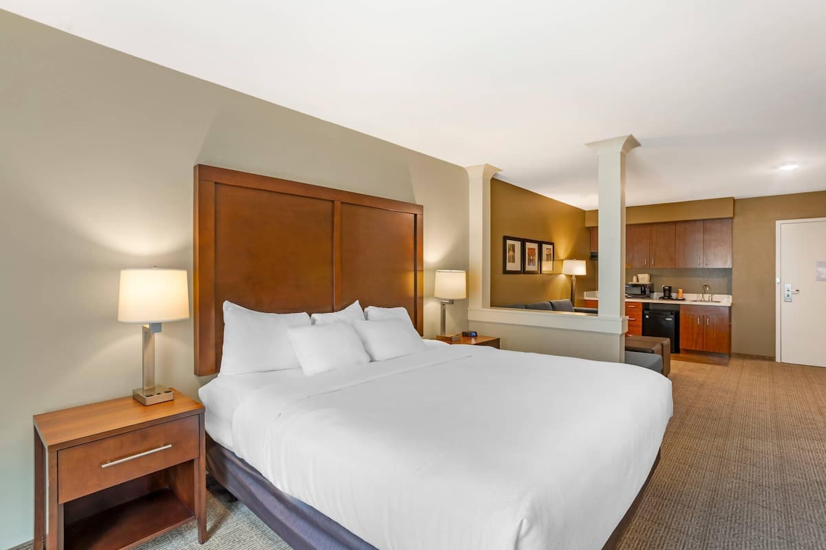 A hotel room with a large bed, a wooden headboard, a nightstand with a lamp, a small seating area, and a kitchenette in the background.