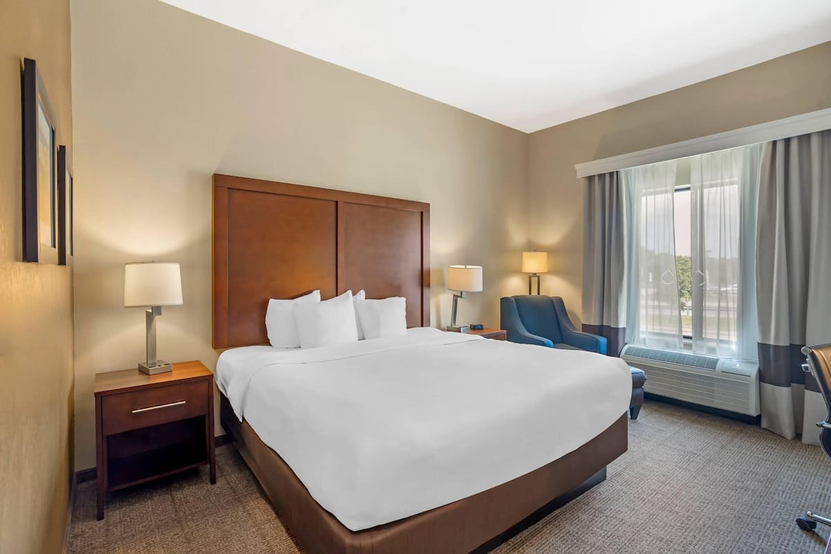 A hotel room with a large bed, two bedside tables with lamps, a chair, and a large window with curtains.