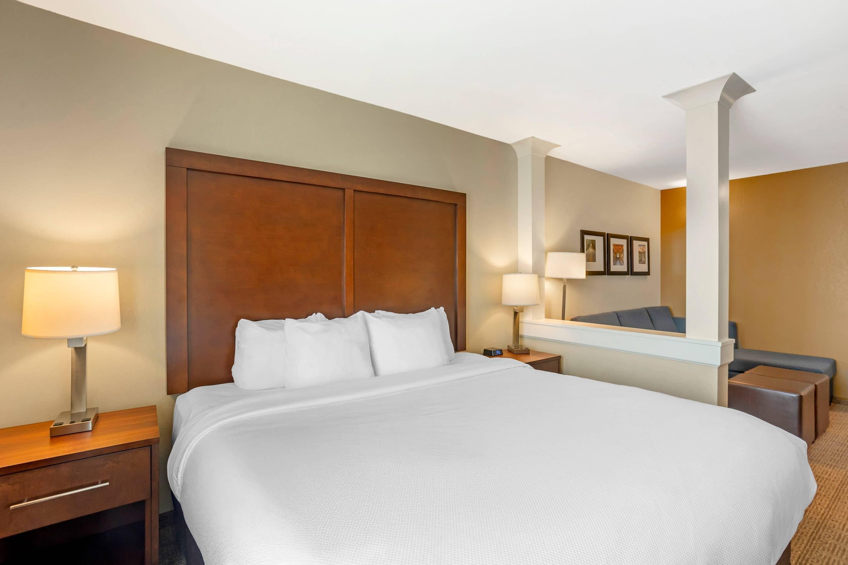 A hotel room with a large bed, a wooden headboard, a nightstand with a lamp, a seating area with a bench, and a wall-mounted mirror.