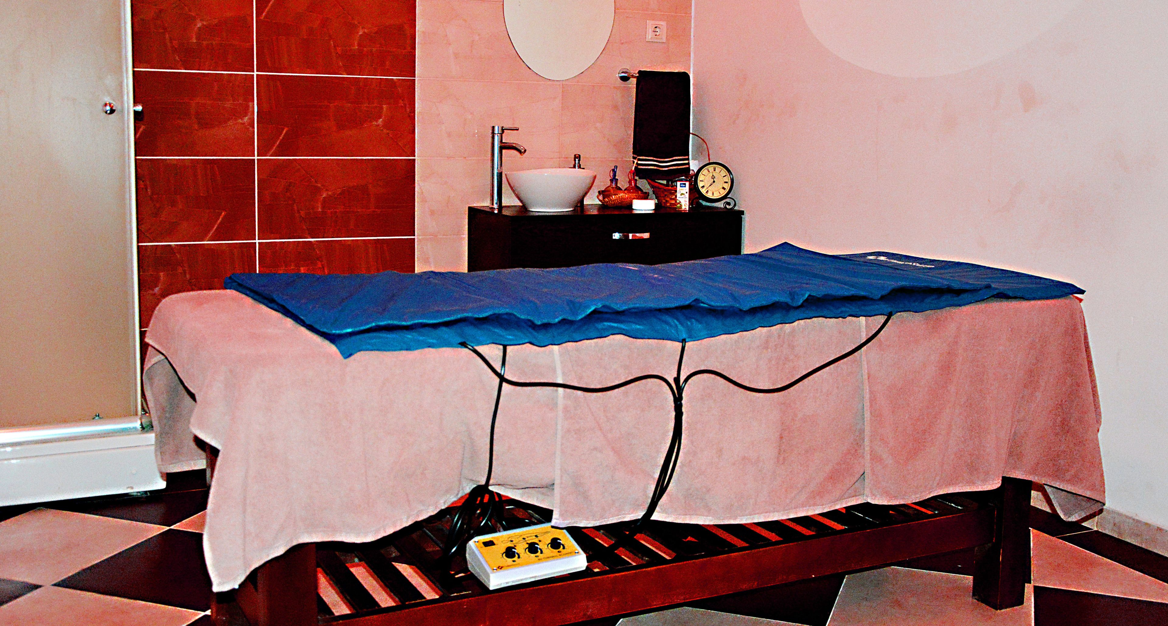 A room with a bed covered by a blue sheet, a small table with a white basin and a clock, and a red-tiled wall.