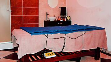 A room with a bed covered by a blue sheet, a small table with a white basin and a clock, and a red-tiled wall.