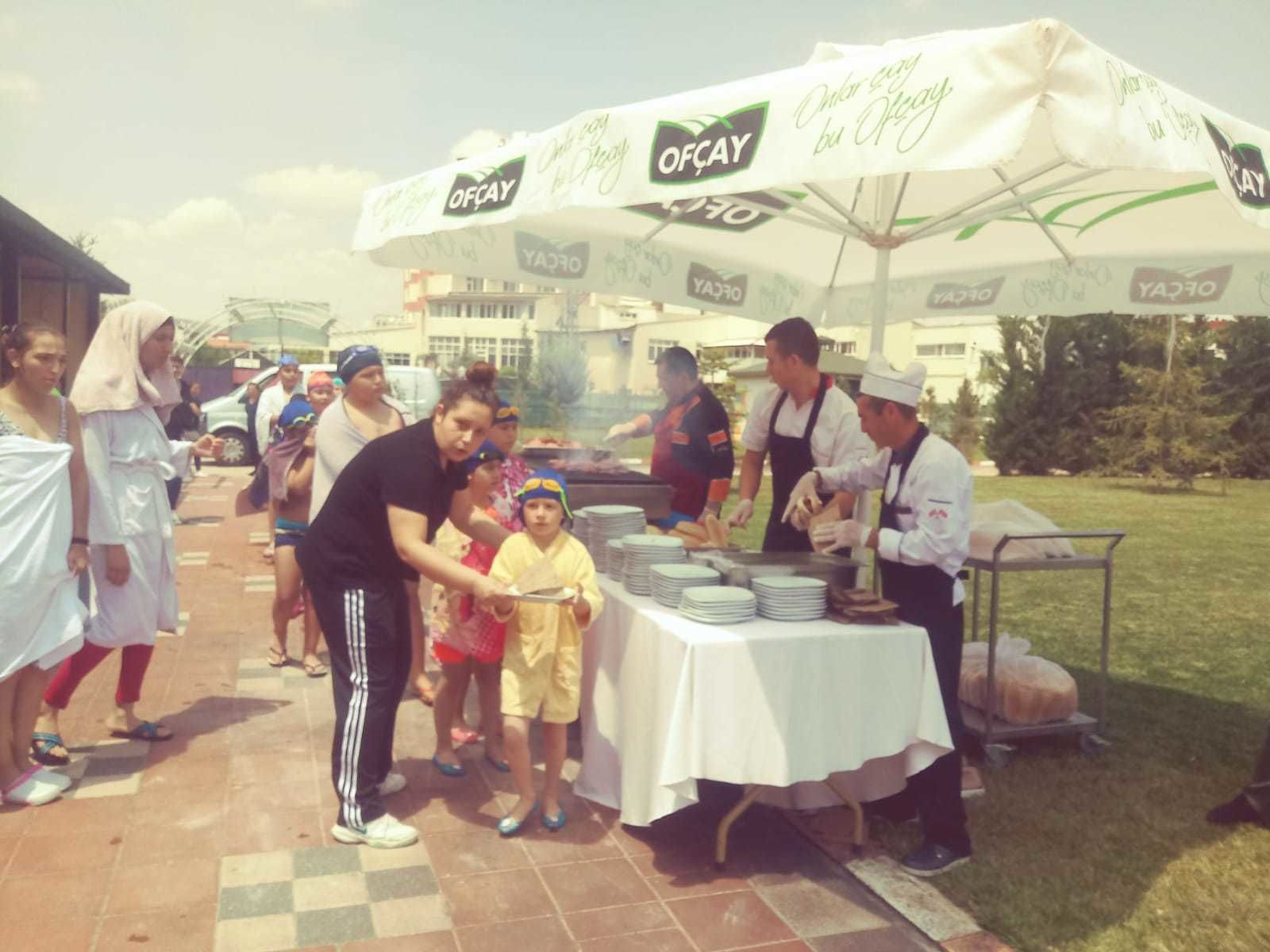 Un grupo de personas en un evento al aire libre, con una mesa cubierta con mantel blanco y un toldo con la marca 'OFCAY'.