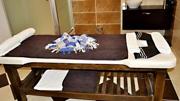A wooden massage table with towels and a blue cloth in a bathroom with a tiled floor and a dark wooden vanity.