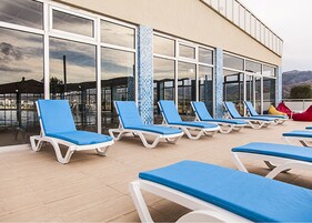 Zona junto a la piscina con varios sillones de descanso y un edificio grande con ventanas reflectantes.
