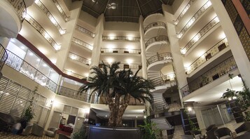 Un vestíbulo de hotel de varios niveles con una palmera central, múltiples balcones con barandillas ornamentadas y un diseño interior moderno.
