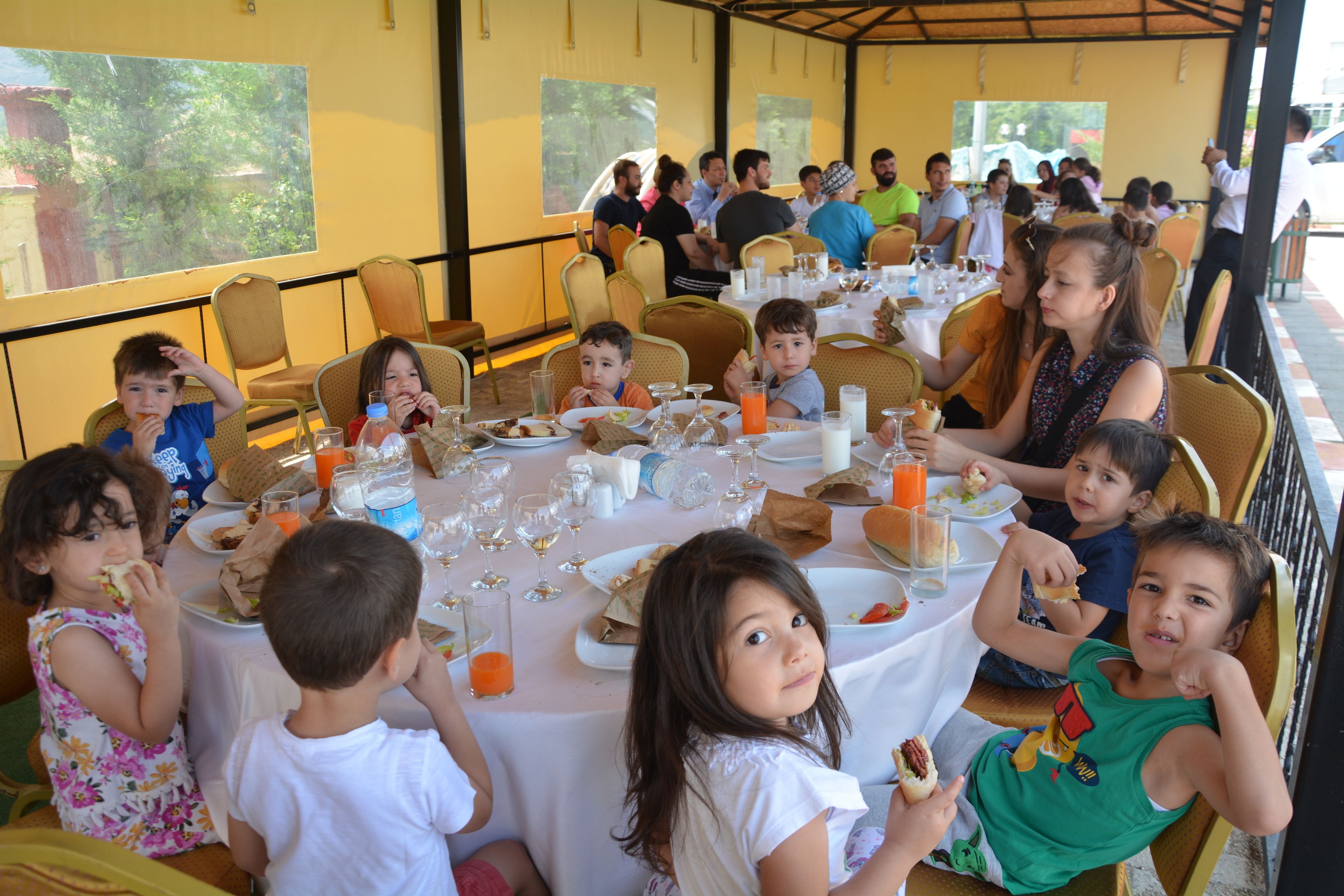 Un grupo de personas sentadas a una mesa con comida y bebidas, bajo una zona cubierta con ventanas.