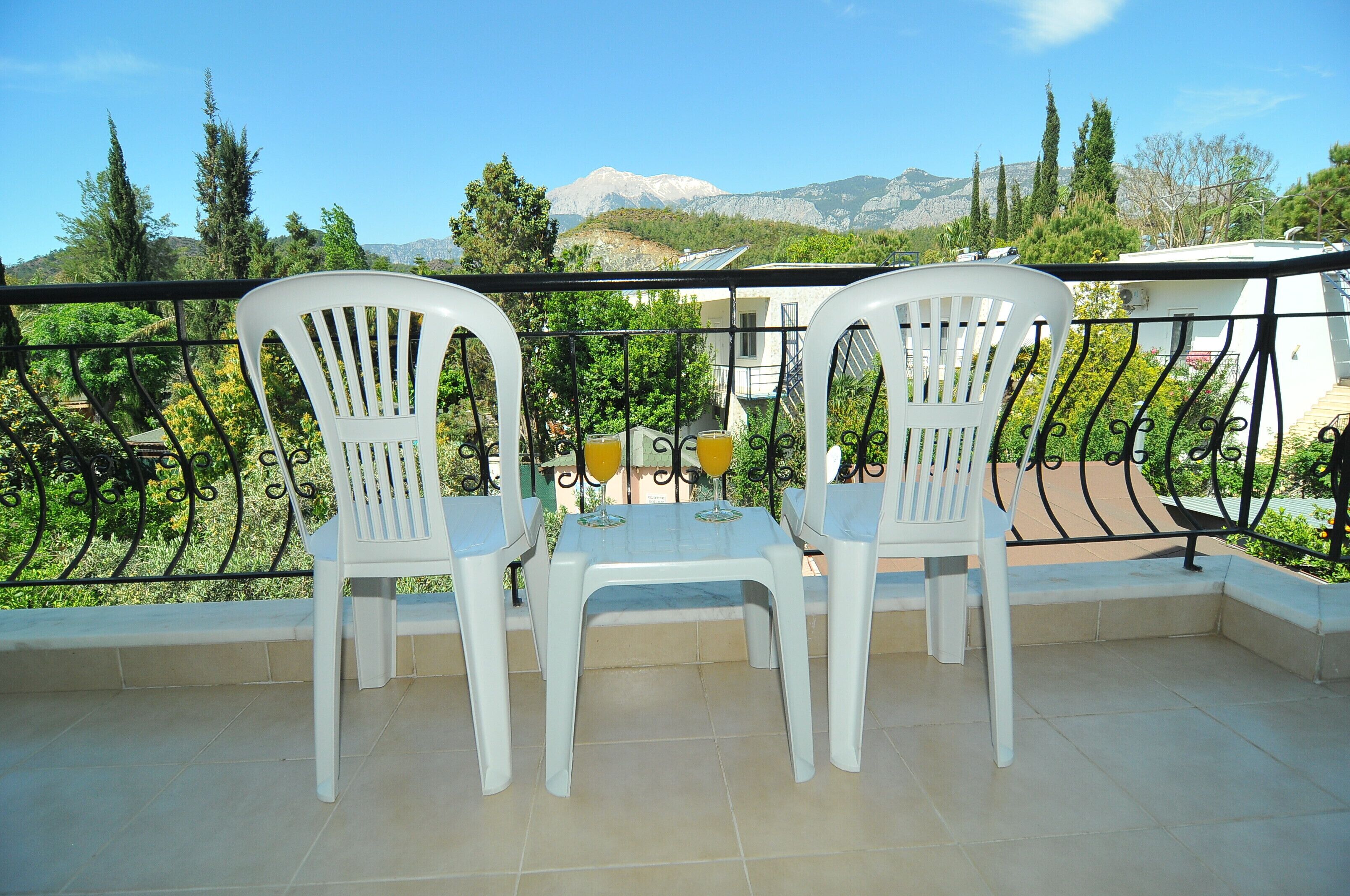 A balcony with two white plastic chairs and a small table, overlooking a mountainous landscape with trees and buildings.