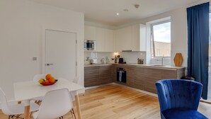 A modern kitchen with white cabinets, a microwave, and a sink. There is a dining table with chairs and a fruit bowl. A blue chair is visible in the foreground. - Premium Hounslow Studio Apartments (Hounslow)