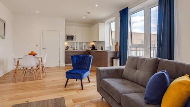 A modern living room with a grey sofa, a blue armchair, and a dining area with a white table and chairs. - Premium Hounslow Studio Apartments (Hounslow)