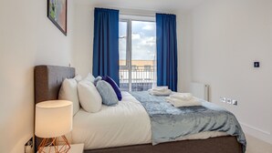 A modern hotel room with a large bed, a bedside lamp, and a view of the outside through a window with blue curtains. - Premium Hounslow Studio Apartments (Hounslow)