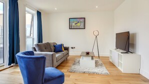 A modern living room with a grey sofa, blue armchair, and a flat-screen TV on a white stand. - Premium Hounslow Studio Apartments (Hounslow)