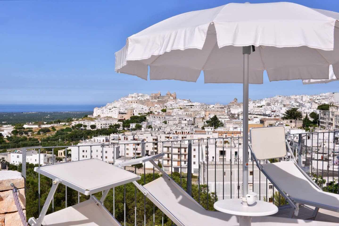 A terrace with white sun loungers and a large umbrella, overlooking a densely packed cityscape with white buildings and a view of the sea.