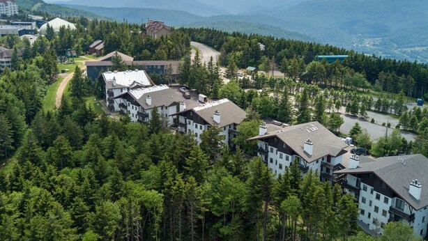 A residential area with multiple buildings surrounded by dense forest and mountains in the background.