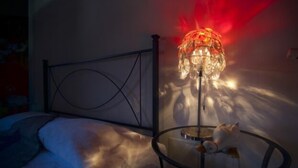 Un lit avec une tête de lit en métal, une table ronde sur laquelle est posé un chat, et une lampe de sol diffusant une lumière rouge.
