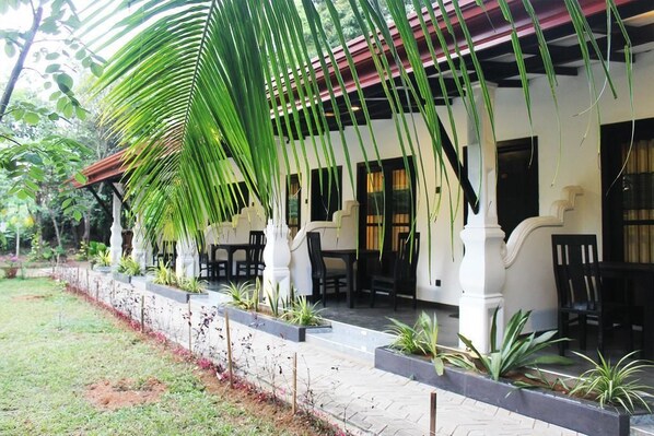 A covered outdoor seating area with white pillars and black chairs, surrounded by potted plants and a brick pathway. - Amron Resort Sigiriya (Sigiriya)