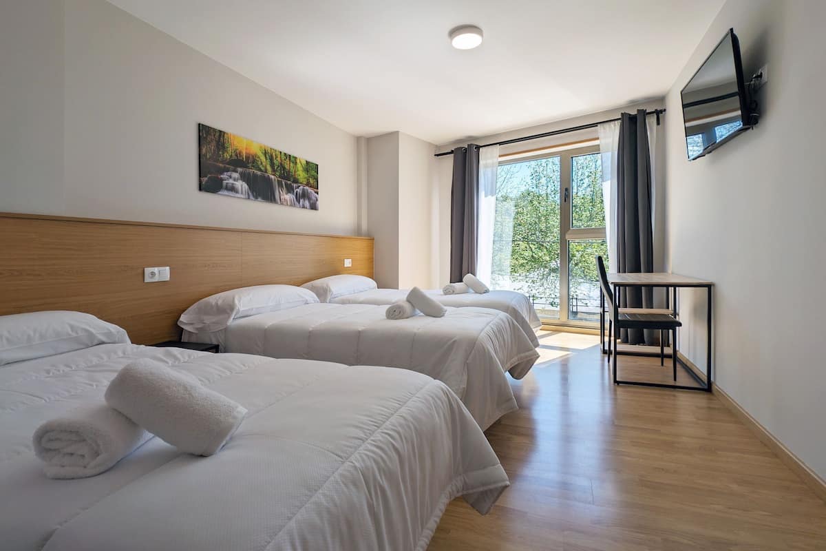 A hotel room with three beds, a desk, a TV, and a window with curtains.