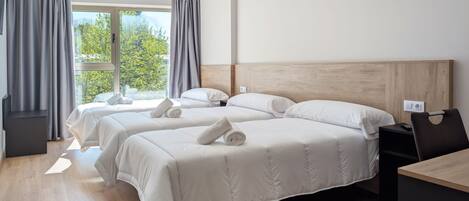 Habitación de hotel con tres camas, cabecero de madera, escritorio y vista a árboles por la ventana.