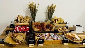A variety of breads, pastries, and condiments on a wooden table with labels indicating types like 'Pan paysés' and 'Galletas'.