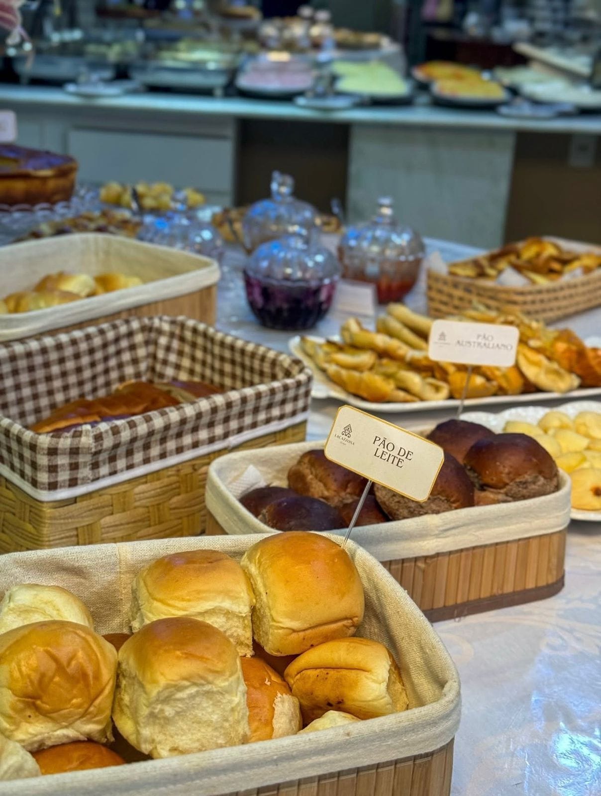 A buffet table with various food items including bread rolls, pastries, and a sign indicating 'Pão de Leite'.
