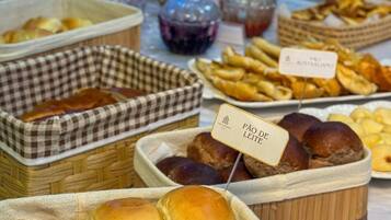 A buffet table with various food items including bread rolls, pastries, and a sign indicating 'Pão de Leite'.