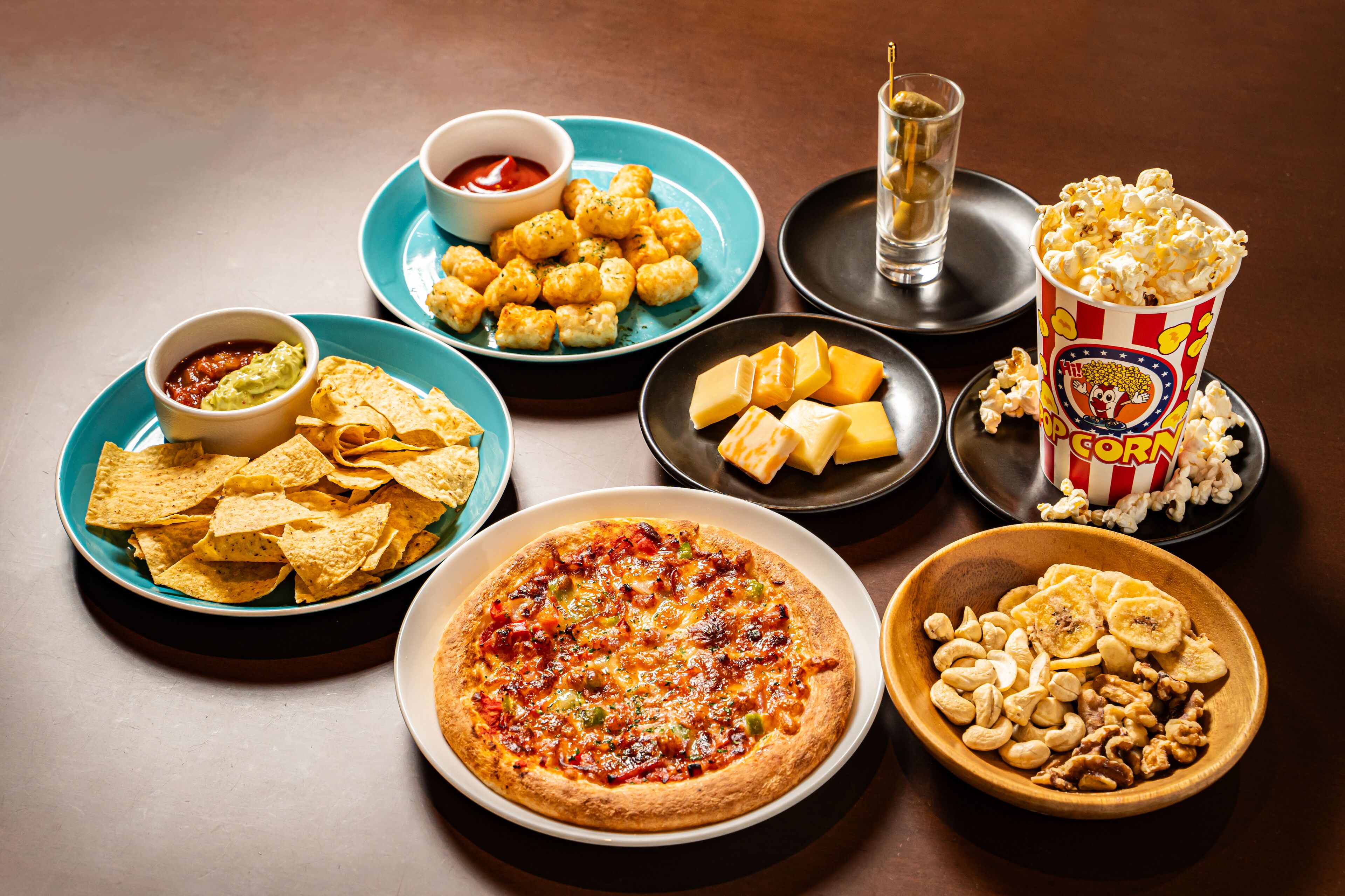 A table with a pizza, nachos, tater tots, popcorn, and a bowl of nuts.