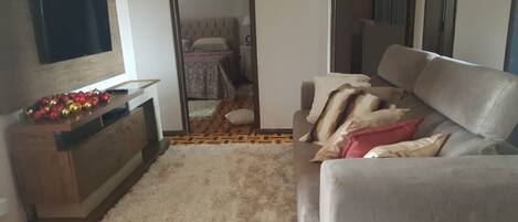 A living room with a grey sofa, a flat-screen TV mounted on the wall, a wooden cabinet with decorative items, and a patterned rug.