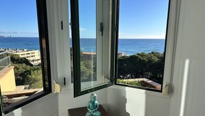 A room with a large window overlooking a coastal view, a small wooden table with a statue, and a white wall.