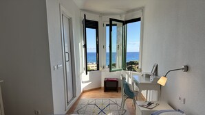 A bedroom with a bed, a desk, a chair, and a view of the sea through open doors.