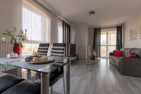 A modern living room with a glass dining table, striped chairs, a grey sofa with red cushions, and a TV.