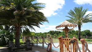 Zona de piscina con sillas de madera, cerca de madera y una palmera.