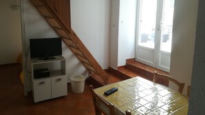 Una habitación con una escalera de madera, un televisor sobre un mueble blanco, una mesa azulejada con un control remoto y una ventana con vistas al exterior.