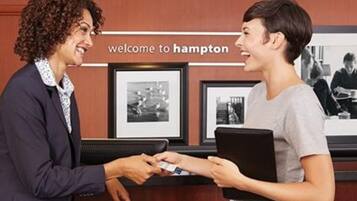 Un mostrador de recepción con un cartel que dice 'welcome to Hampton'. Hay fotografías enmarcadas en la pared detrás del mostrador.