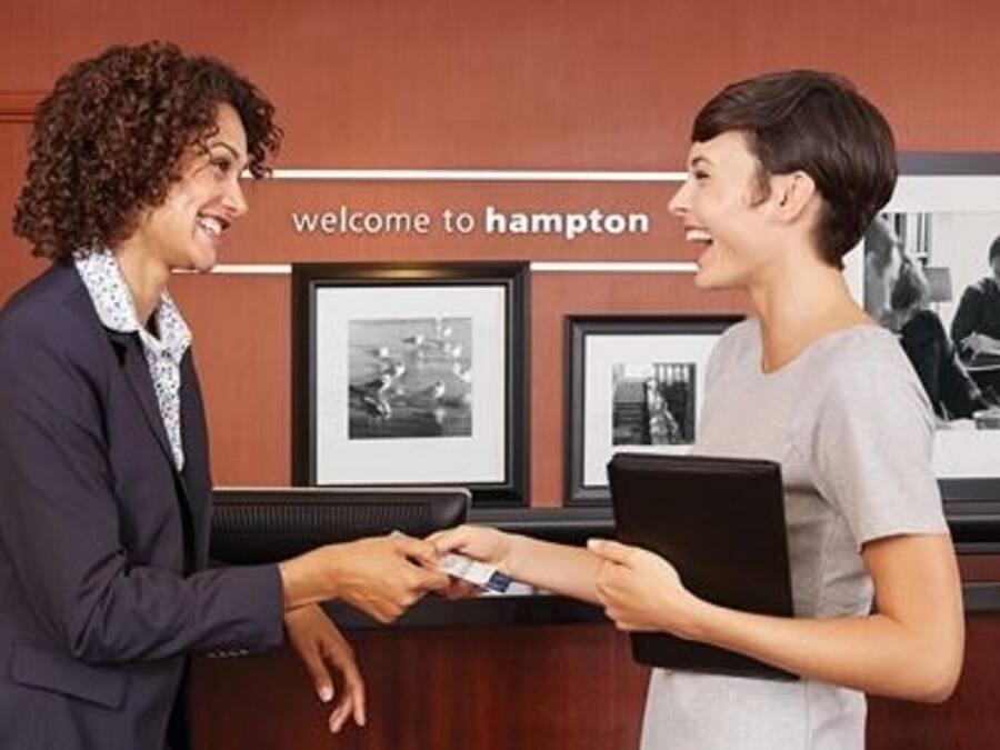 Un mostrador de recepción con un cartel que dice 'welcome to Hampton'. Hay fotografías enmarcadas en la pared detrás del mostrador.