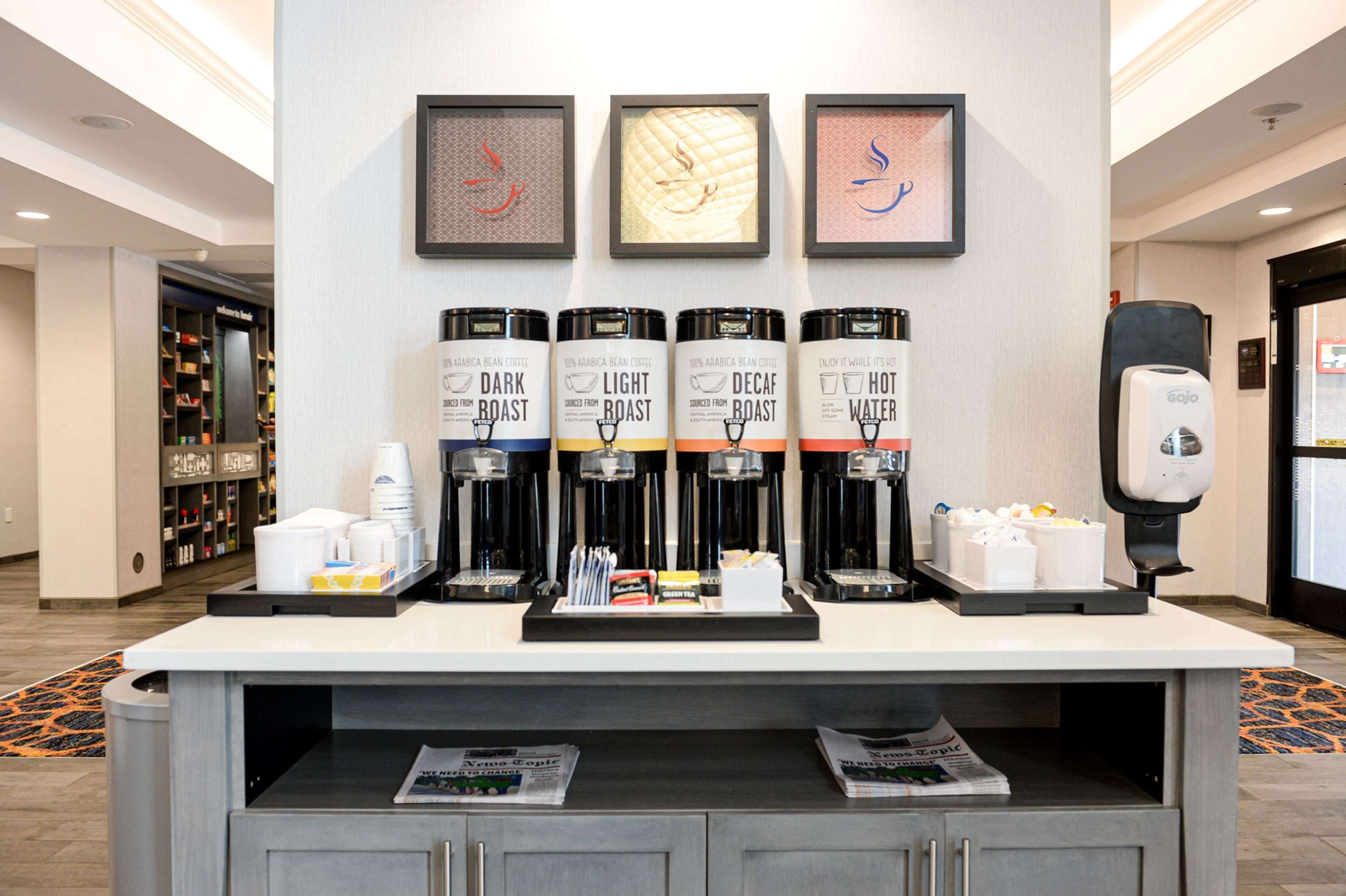 Una estació de cafè amb quatre dosificadors de cafè etiquetats 'Dark Roast', 'Light Roast', 'Decaf' i 'Aigua Calenta'. Hi ha gots, tovalloles i un dispensador de sabó per mans al taulell.