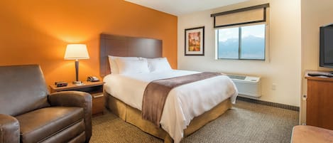 A hotel room with a bed, a chair, a lamp, a nightstand, a TV, and a window with a mountain view.