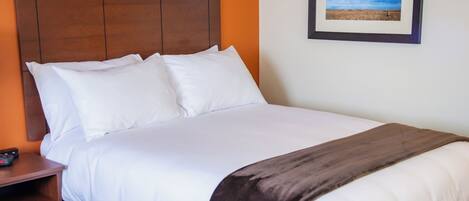 A neatly made bed with white linens and a brown throw blanket, a wooden nightstand with a remote control, and a framed picture of a landscape on the wall.